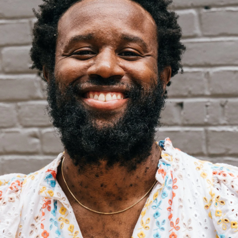 Artist Jerome Ellis smiling and wearing a white dress with colorful floral patterns