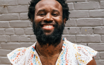 Artist Jerome Ellis smiling and wearing a white dress with colorful floral patterns